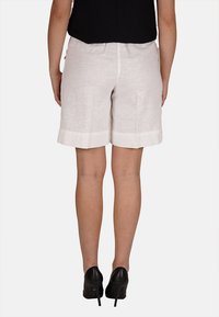 Pantaloni corti in lino di colore beige chiaro con orlo risvoltato. La vista posteriore mostra una texture senza pieghe e una vestibilità comoda. Si indossano tacchi alti neri.