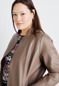Chaqueta de cuero marrón con cuello alto, textura suave y cierre de cremallera, llevada sobre una camiseta negra con un diseño de leopardo.