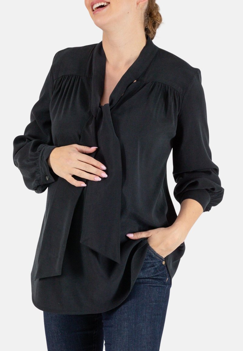 Schwarze Bluse mit einem Bindekragen, ausgestattet mit Falten an den Schultern und langen Ärmeln, aus leichtem, glattem Stoff. Getragen mit blauen Jeans.
