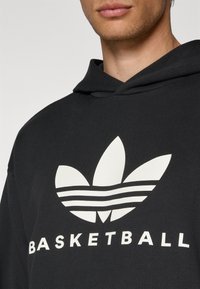 Fekete kapucnis felső, nagy fehér Adidas logóval, amely három levelet és vízszintes csíkokat tartalmaz, az "BASKETBALL" szóval nyomtatva alatta.