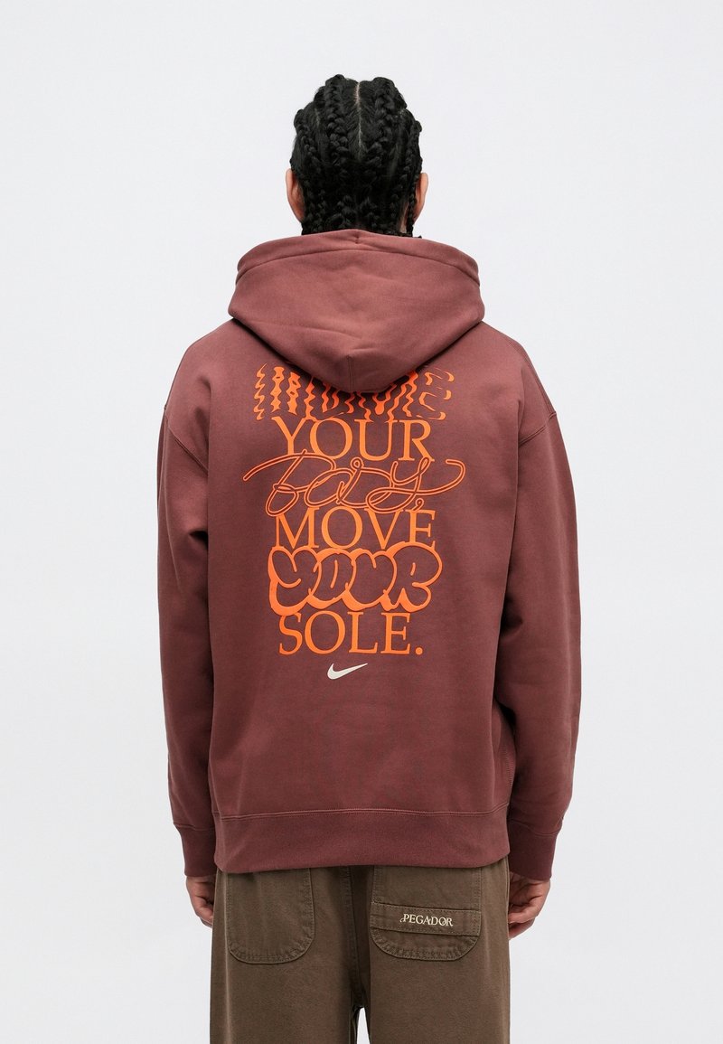 Personne aux cheveux tressés portant un sweat à capuche bordeaux avec un texte orange et un petit logo Nike dans le dos, ainsi qu’un pantalon marron avec un écusson.