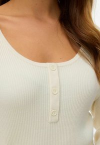 Top in crema a coste con scollo tondo e tre bottoni sul davanti. Il tessuto è morbido con una superficie leggermente strutturata.