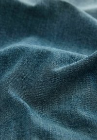 Denim-kangas syvässä sinisessä sävyssä, jossa on sileä rakenne näkyvine kudontakuvioineen ja kevyt kiilto taitteissa.