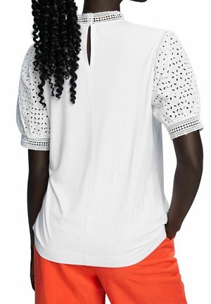 Bílý top s vyvýšenými rukávy z broderie anglaise, detaily na zádech ve tvaru klíče a krajkovým lemem, v kombinaci s oranžovými spodními částmi, které mají uvolněný střih.