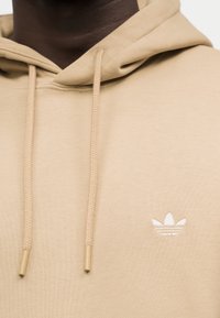 Μπεζ φούτερ με κορδόνια και μικρό λογότυπο Adidas Trefoil σε λευκό στο στήθος, φορεμένο από άτομο με σκούρο δέρμα.
