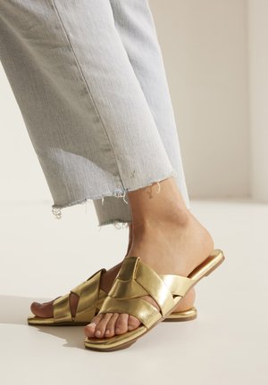 Goldene Leder-Slidesandalen mit einem Kreuzdesign, flacher Sohle und offener Zehenpartie. Getragen mit hellblauen, zerfetzten Jeans.