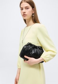 Νεαρή γυναίκα με μακρυμάνικο φόρεμα ανοιχτό κίτρινο κρατάει μεγάλο μαύρο τσάντα φάκελο με πιέτες, φοράει σκουλαρίκια με κρεμαστά μαργαριτάρια και μακρύ κολιέ.