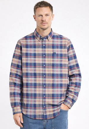 Homme portant une chemise à manches longues à rayures carreaux dans les tons bleus, beiges et rouges, une main dans la poche, sur fond blanc uni.