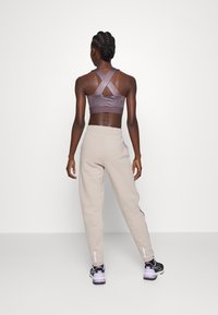 Szary sportowy biustonosz z krzyżującymi się paskami i logo, w połączeniu z beżowymi spodniami dresowymi z bocznymi kieszeniami i odblaskowymi akcentami. Czarne buty sportowe.