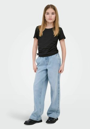 Ung pige stående iført sort kortærmet top, løse lyseblå jeans og sorte sko, foran en ensfarvet lys baggrund.