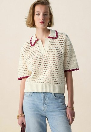 Jonge vrouw die een crèmekleurige, korte mouws trui van crochet draagt met bordeauxrode afwerking en lichtblauwe spijkerbroek, staand tegen een beige achtergrond.