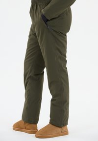 Pantalons légers et isolés, de couleur vert olive, avec une taille élastique et des poches latérales. Portés avec des chaussons en suède marron clair.