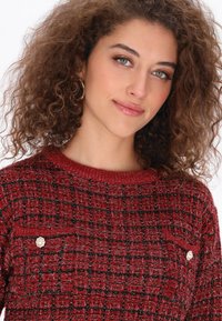 Czerwony sweter o teksturowanej powierzchni z plecionym wzorem, okrągłym dekoltem i metalowymi guzikami na kieszeniach na piersi.