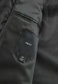 Schwarzer Stoff mit einer subtilen gerippten Textur; enthält ein Etikett mit "NEXT", Größe "M" und "Hergestellt in China", das an der Seite angenäht ist.