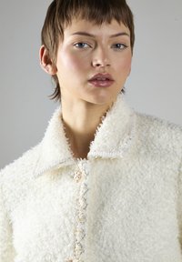 Hvid tekstureret jakke med en stor krave, dekorativ syning og centrallukning med knap. Blødt, fluffy materiale med et hyggeligt udseende.