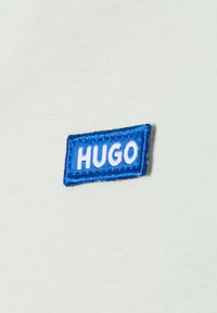 Jasnozielona bawełniana koszulka z prostokątną niebieską haftowaną naszywką z białym napisem "HUGO". Gładka struktura materiału.