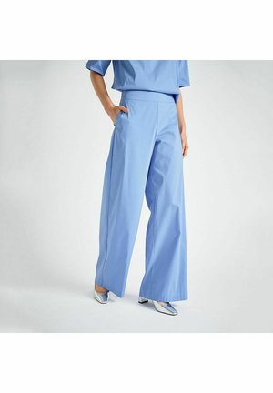 Vrouw die lichtblauwe, wijd uitlopende broek en bijpassende top draagt, staand met één hand in de zak, draagt zilverkleurige schoenen met blokhak.