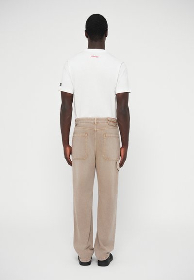 Λευκό T-shirt με κόκκινο λογότυπο στην πλάτη και μπεζ παντελόνι φαρδιάς γραμμής με δύο πίσω τσέπες και πλαϊνή τσέπη.