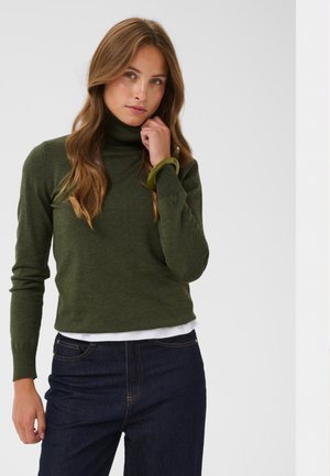 Mujer con cabello largo y ondulado que lleva un suéter de cuello alto verde oliva, vaqueros azul oscuro y una pulsera verde, de pie frente a un fondo ligero y liso.