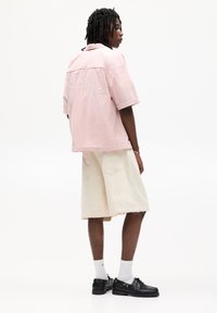 Camisetta rosa a maniche corte con ricamo "Sunset Chase" sul retro, abbinata a pantaloni corti color crema e scarpe nere. Design semplice e casual.