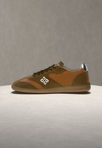 Baskets basses marron et orange avec des lacets brun foncé et un logo blanc sur le côté, exposées sur une surface claire avec un fond gris.