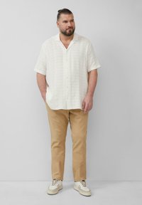 Mann mit Bart und zurückgebundenem Haar steht in einem kurzärmligen weißen Hemd, beigen Hosen und weißen Turnschuhen vor einem schlichten Hintergrund.