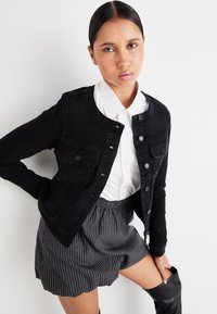Μαύρο τζάκετ από τζιν με στρογγυλή λαιμόκοψη, δύο τσέπες στο στήθος και ασημένια κουμπιά, συνδυασμένο με λευκό πουκάμισο με γιακά και ριγέ σορτς.