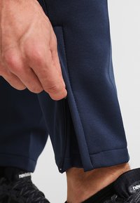 Marineblå træningsbukser med en lynlåsdetaile nær kanten. Stoffet har en struktureret overflade. Båret med sorte sneakers.