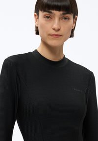 Kobieta o krótkich ciemnych włosach, ubrana w obcisłą czarną bluzkę z długim rękawem o teksturze i subtelnych poziomych paskach, na białym tle.