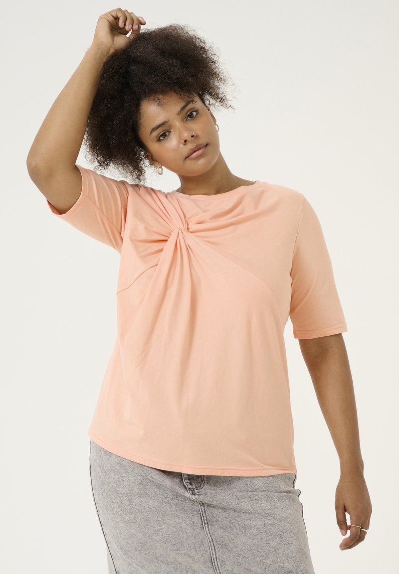Peachfarvet, kortærmet T-shirt med en snoet detalje foran, parret med en lysegrå denimnederdel. Glat stoffets tekstur.