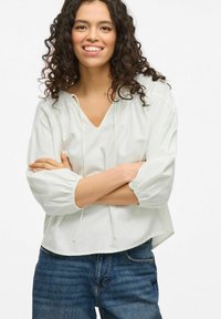 Hvit, lett bluse med avslappet passform, V-hals og snor. Har utsvungne ermer og en cropped passform. Kombinert med mørkeblå jeans.