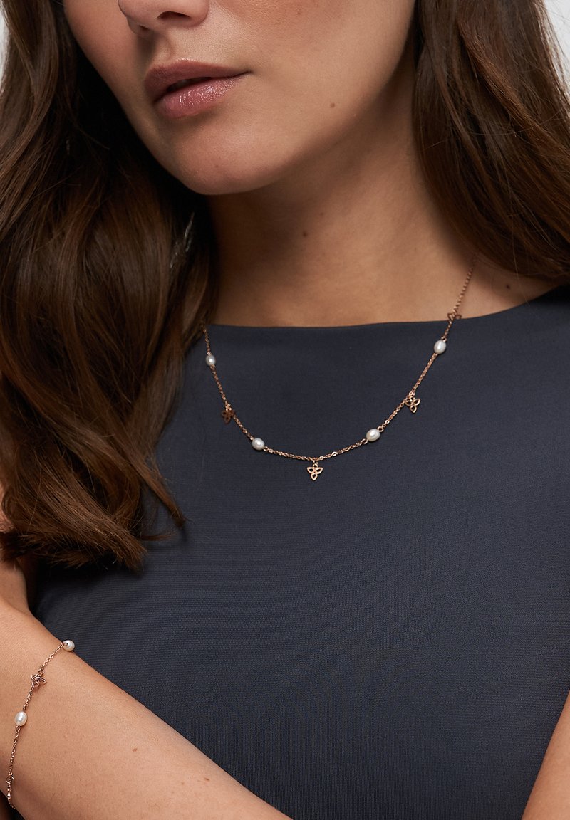 Roségouden ketting met parels en decoratieve accenten, met een fijn kettinkje ontwerp. Model draagt het om de hals, met een parelarmband zichtbaar.
