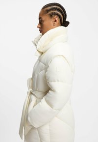 Vit pufferjacka med hög krage, midjebälte och ribbade detaljer. Tyget ser slätt och något glänsande ut, med en quiltad design.