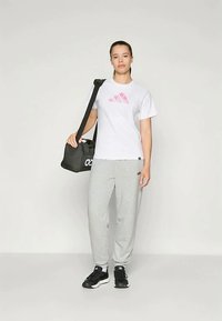 Bijela majica s ružičastim logotipom, sivi trenirke, nosi crni torbu s bjelim brendingom i ima crne sportske cipele.