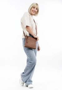 Borsa a tracolla in pelle marrone con una texture liscia, forma rettangolare e dettagli con zip. Il modello indossa un morbido maglione beige e jeans blu.