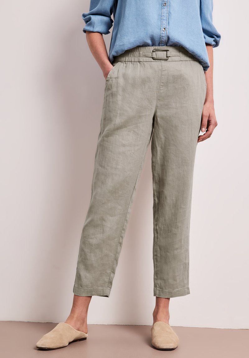 Personne portant une chemise bleu clair à manches longues, un pantalon en lin beige taille haute avec ceinture, et des chaussures beige sans lacets, debout contre un mur crème.