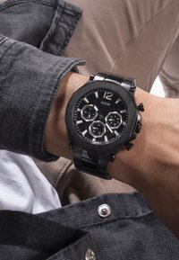 Czarny zegarek Guess z ośmiokątnymi tarczami chronografu, czarnym metalowym paskiem i wyraźnymi białymi znacznikami godzin. Noszony na nadgarstku z szarym materiałem.