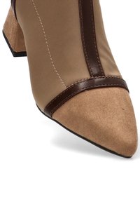 Botín beige de tobillo con punta afilada, acentos de cuero marrón, acabado de ante texturizado y tacón cuadrado. Presenta un detalle de costura contrastante.