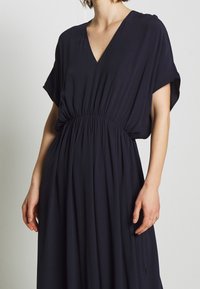 Marineblå kjole med V-hals, korte ærmer, elastisk talje og flydende stof. Har en subtil struktur og en afslappet pasform.