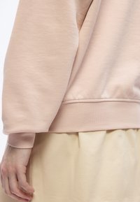 Χέρι ατόμου χαλαρό στο πλευρό, φορούν ανοιχτό ροζ φούτερ με μανσέτα και μπεζ παντελόνι, με φόντο απλό λευκό.