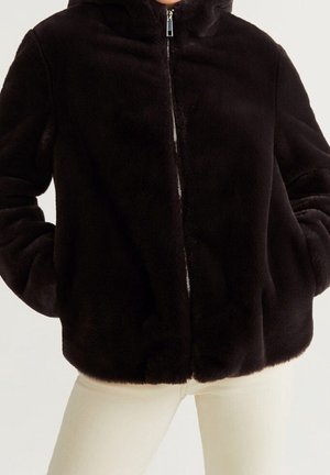 Personne portant une veste zippée noire et duveteuse, mains dans les poches, et un pantalon clair sur un fond uni.