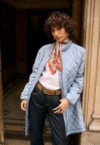 Naine toetub kivisest ukseraamistikule, kandes helesinist tikitud jope, mustrilist salli, valget pluusi, tumedaid pükse ja pruuni vööd.