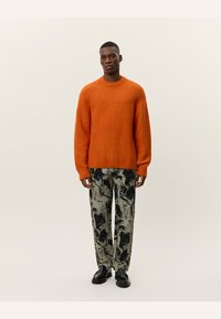 Suéter de lana naranja brillante con cuello redondo, combinado con pantalones de patrón en negro y tonos apagados, y zapatos negros gruesos.