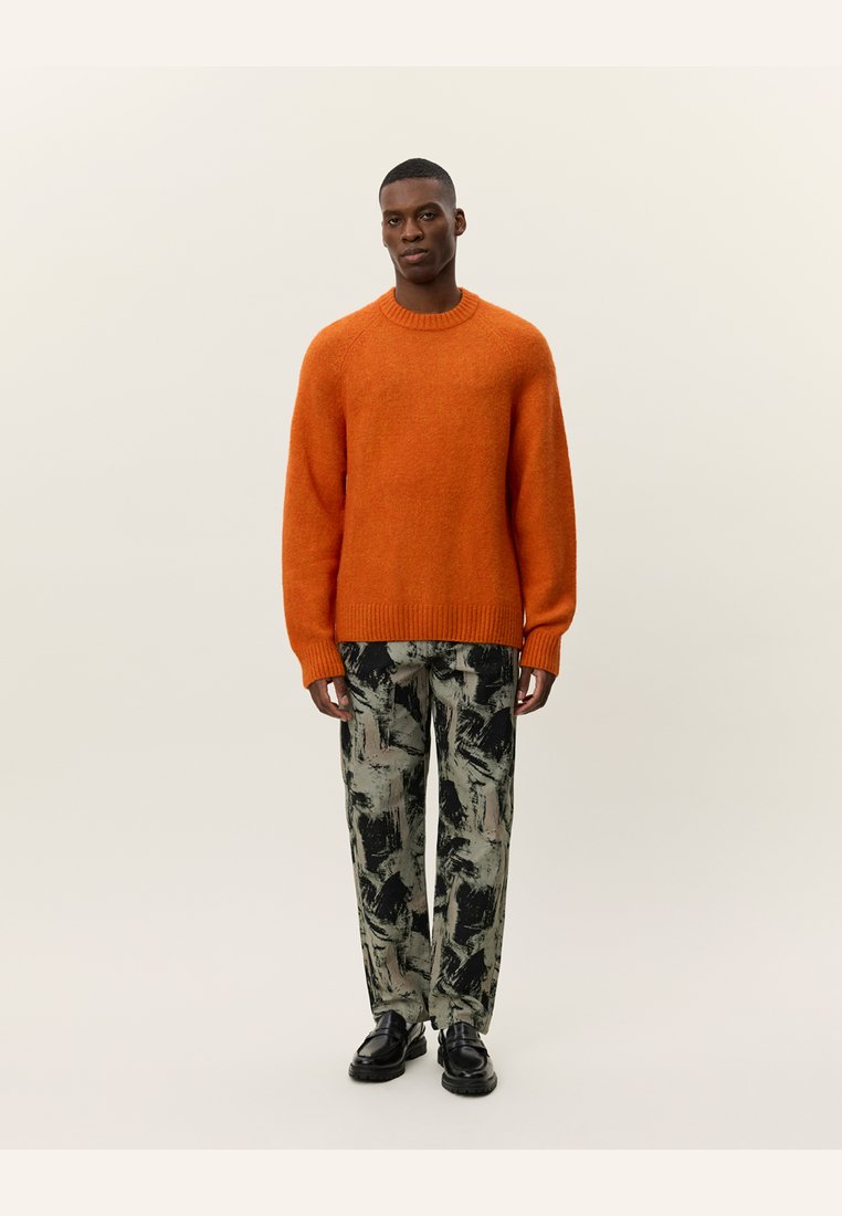 Suéter de lana naranja brillante con cuello redondo, combinado con pantalones de patrón en negro y tonos apagados, y zapatos negros gruesos.
