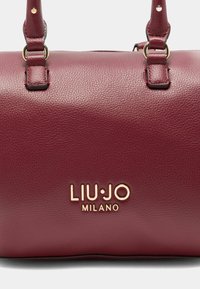 Burgunderfarbene Lederhandtasche mit goldenem Metallzubehör. Verfügt über doppelte Griffe, strukturierte Oberfläche und geprägtes Logo "LIU·JO MILANO" auf der Vorderseite.