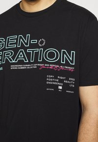 Černé bavlněné tričko se výrazným tyrkysovým a bílým textem, na kterém je výrazně napsáno "GENERATION", a dalšími brandingovými detaily níže.