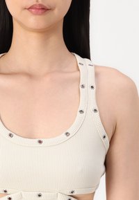 Crop top beige a costine con spalline larghe, caratterizzato da dettagli in occhielli metallici lungo il collo e la parte centrale. Texture liscia e design aderente.