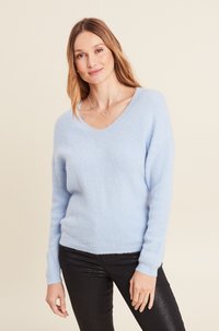 Maglione azzurro chiaro a V realizzato in materiale morbido con maniche lunghe. Indossato con pantaloni neri aderenti e leggermente lucidi.
