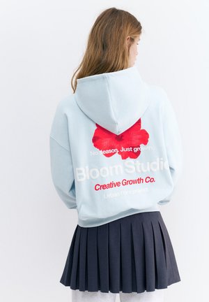 Jeune femme aux longs cheveux portant un sweat à capuche bleu clair avec une fleur rouge et un texte, ainsi qu'une jupe plissée foncée, vue de dos sur fond blanc.