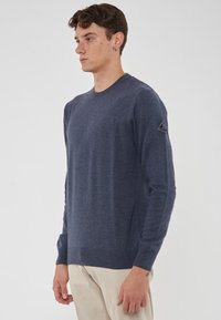 Maglione lavorato a maglia blu navy con scollo a girocollo, dotato di maniche lunghe e una finitura testurizzata. Include un patch triangolare con logo sulla manica.